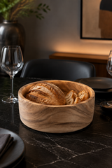 A round wooden bowl filled with rustic bread sits on a dark marble dining table, surrounded by plates, wine glasses, and chairs in a softly lit interior setting.