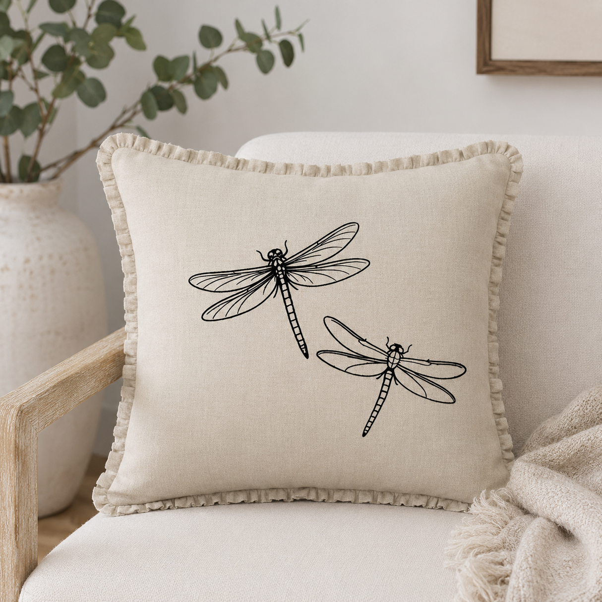 Natural linen cushion cover with black dragonfly print and ruffle edge, styled on a cream armchair with neutral textiles.
