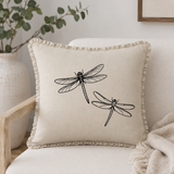 Natural linen cushion cover with black dragonfly print and ruffle edge, styled on a cream armchair with neutral textiles.