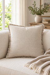 A square beige throw pillow with a delicate ruffled trim, styled on a light-colored sofa next to a matching fringed throw blanket in a bright, neutral-toned living room.
