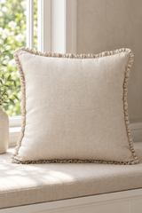 A square beige linen throw pillow with a ruffled trim, resting on a matching beige upholstered window seat in front of a bright window.
