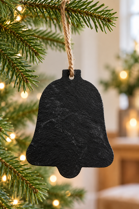 A black slate Christmas bell ornament hanging from a natural jute twine on a green pine tree branch with warm white string lights in the background.
