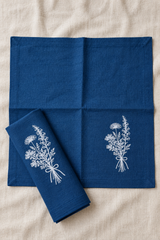Navy Florilegium linen napkins set on a beige fabric background, featuring white embroidered wildflower bouquets. One napkin is laid flat, and another is folded over to show the embroidered detail.