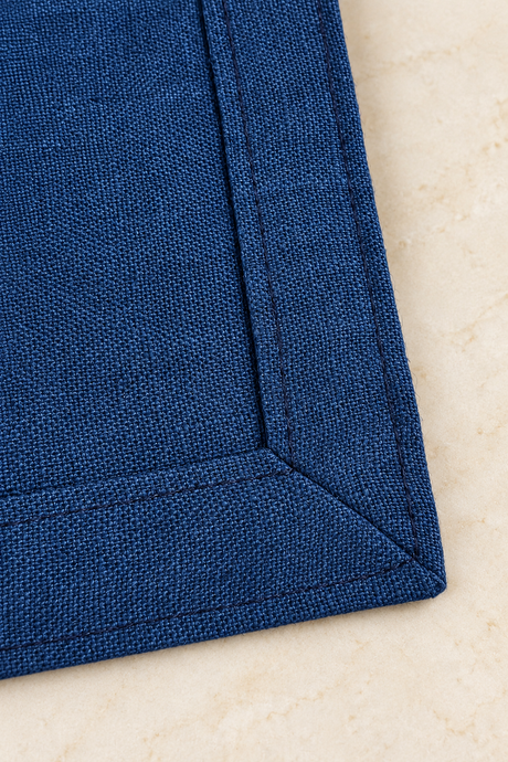 Close-up of the reverse of a navy linen napkin, showing its 2.5cm double-fold hem, matching stitching and mitred corner.
