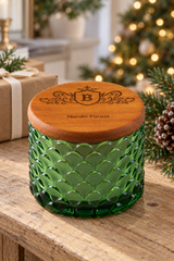 A green textured glass jar candle with a round wooden lid engraved with a monogram and the words 'Nordic Forest,' shown on a wooden table with a gift box and Christmas lights in the background.