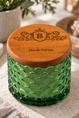 A green textured glass jar with a round wooden lid engraved with a monogram and the words 'Nordic Forest', shown on a neutral stone surface with soft-focus background decor.
