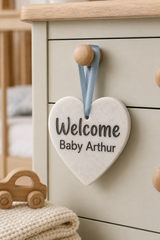 A heart-shaped personalised baby shower decoration hanging on a cream coloured dresser. The stone plaque reads 'Welcome Baby Arthur' in dark lettering and is suspended by a dusty blue ribbon with wooden beads.