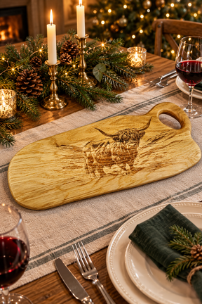 A rectangular wooden serving board with a handle, featuring a engraved Highland cow design, displayed on a decorated holiday dinner table with candles, pinecones, wine glasses, and plates.