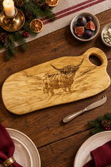 A wooden serving board with a carved Highland cow design, styled on a rustic wooden table with a knife, plates, figs, and pine branches.