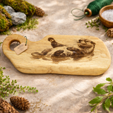 Wooden cutting board with engraved design of otter on a natural stone surface with greenery and pinecones.