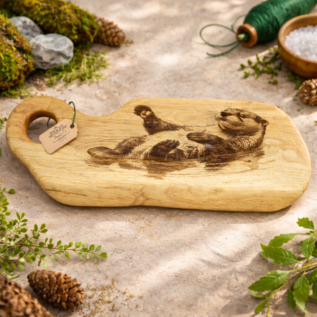 Wooden cutting board with engraved design of otter on a natural stone surface with greenery and pinecones.
