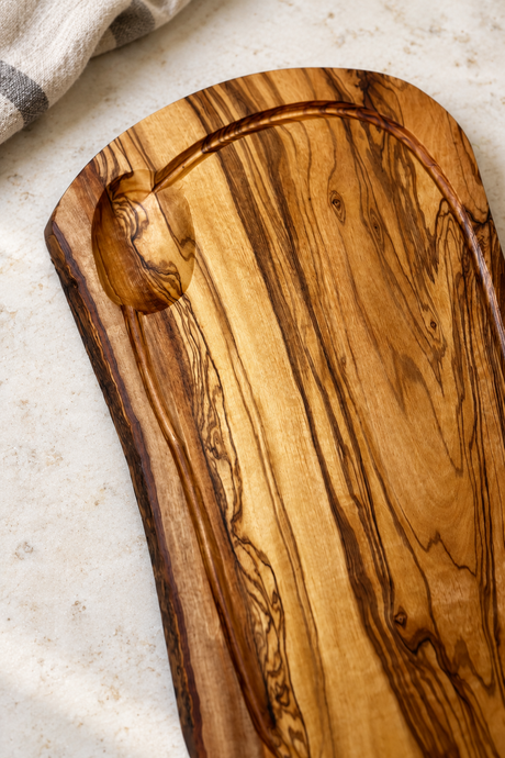 An olive wood serving board with a natural live edge and a carved recessed area, shown on a light textured surface with a folded cloth in the corner.
