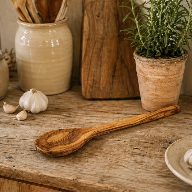 Wooden spoon on a wooden surface with a potted plant and garlic in the background.