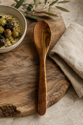 Olive Wood spoon on a wooden cutting board with a bowl of food and a napkin in the background.