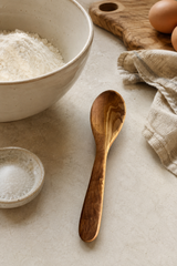 Olive Wood spoon on a light surface with a bowl of flour, eggs, and a cloth in the background.