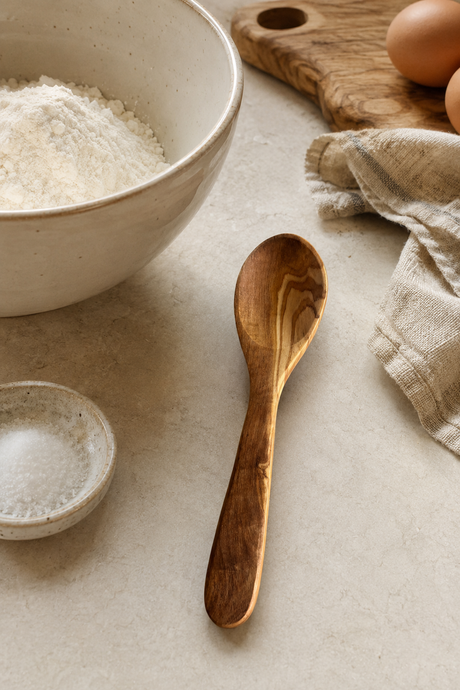 Olive Wood spoon on a light surface with a bowl of flour, eggs, and a cloth in the background.
