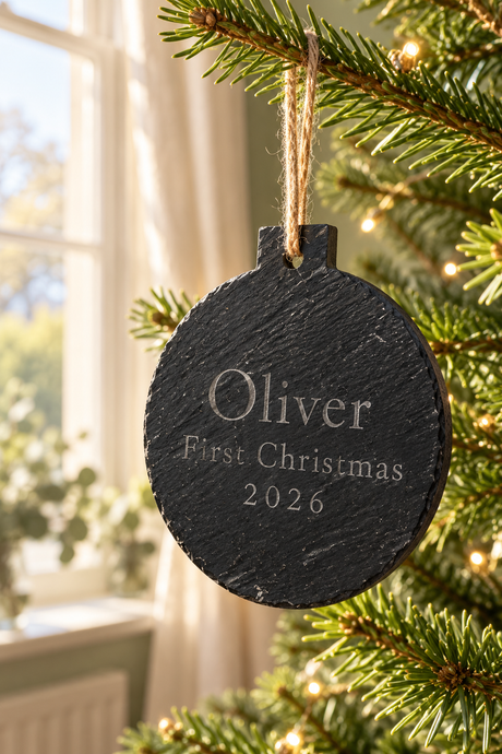 A round black slate Christmas ornament engraved with 'Oliver First Christmas 2026' hanging from a pine tree branch with warm string lights in the background.