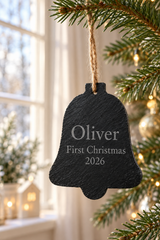 A personalised black slate Christmas bell ornament hanging from a tree branch with a twine loop. The bell is engraved with the name 'Oliver', 'First Christmas', and '2026' in white text.