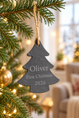 A personalised Christmas tree ornament shaped like a pine tree, made of dark slate with engraved text reading 'Oliver First Christmas 2026', hanging from a natural jute string on a decorated Christmas tree branch in a cozy living room setting.