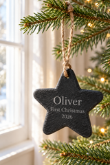 A personalized black slate Christmas star ornament hanging from a pine tree branch with a twine loop. The star is engraved with the name 'Oliver', 'First Christmas', and '2026' in white lettering.