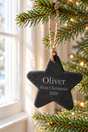 A personalized black slate Christmas star ornament hanging from a pine tree branch with a twine loop. The star is engraved with the name 'Oliver', 'First Christmas', and '2026' in white lettering.