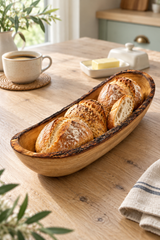 A rustic wooden boat-shaped bread bowl filled with freshly baked sourdough bread rolls, set on a wooden dining table in a warm, softly lit kitchen setting.