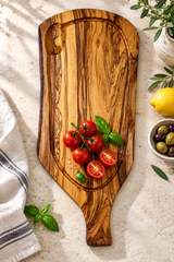 An olive wood serving board with a handle, styled with fresh tomatoes, basil, a lemon, and olives on a textured light beige surface.