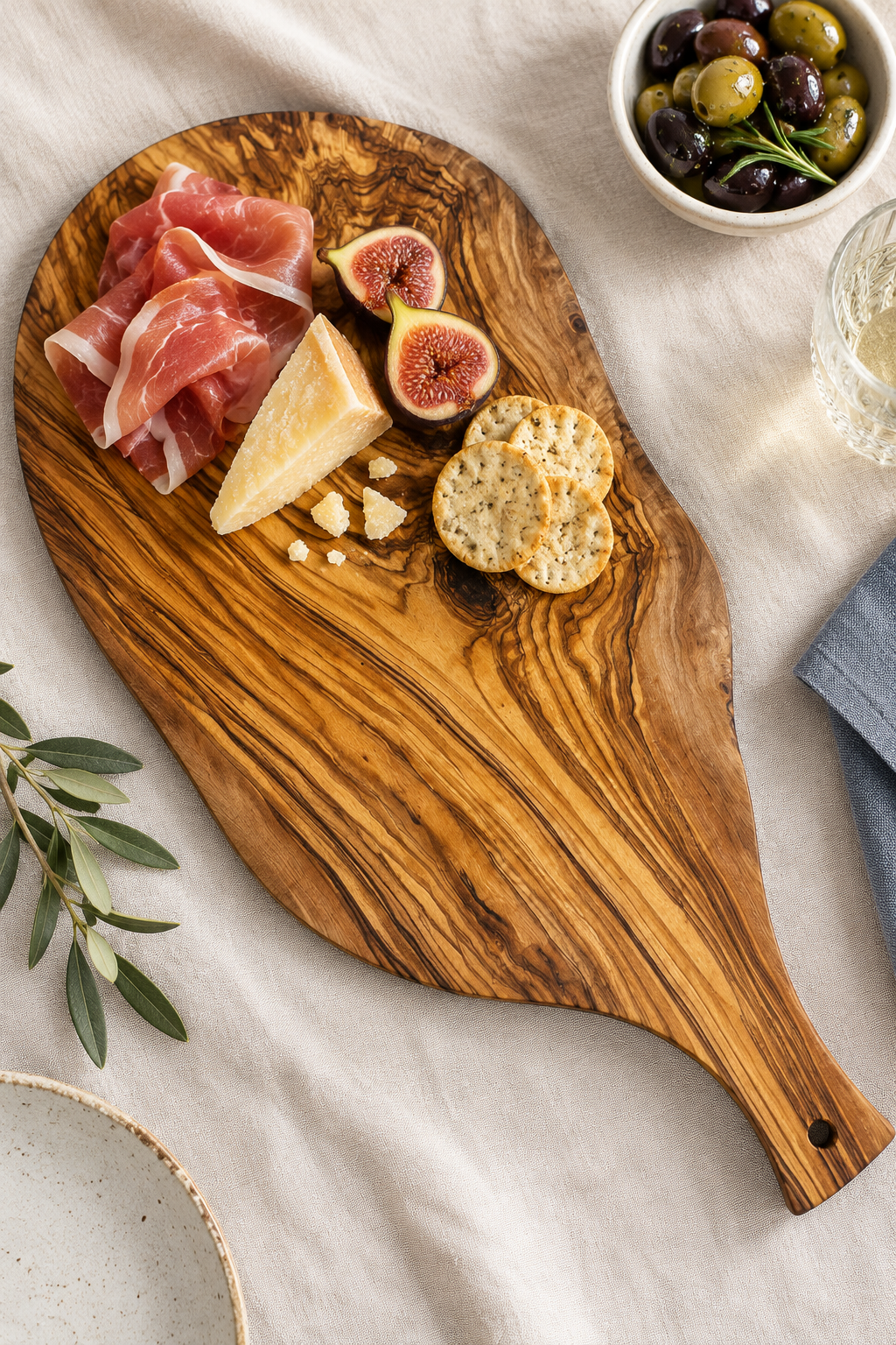 An olive wood serving board with a handle, styled with food including sliced prosciutto, cheese, figs, crackers, and olives on a light linen tablecloth.