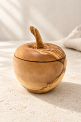 A handcrafted olive wood container shaped like an apple, featuring a matching wooden lid with a stem detail, set on a textured light beige surface.