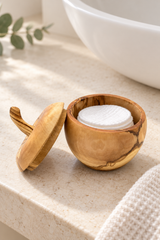 A small round wooden container with a matching lid, shown open on a light stone countertop. Inside the container are white round cotton pads, with a white bowl and a folded cream towel in the softly blurred background.