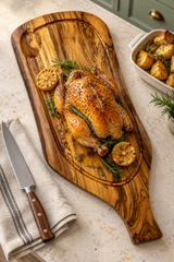 An olive wood carving board with a rustic hand-painted design, styled with a roasted chicken, lemon slices, rosemary, and a chef's knife on a light beige marble countertop.
