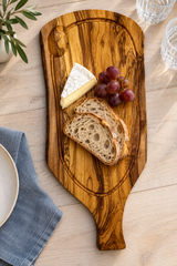 An olive wood serving board with a handle, styled on a light wood tabletop with sliced bread, a wedge of brie cheese, and red grapes.