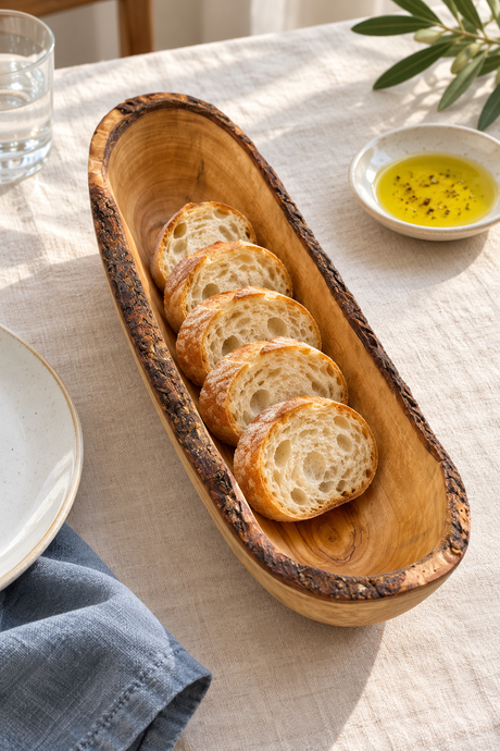 A long oval wooden serving bowl with a dark bark-like rim, filled with sliced rustic bread on a light linen tablecloth. A small dish of olive oil, a glass of water, and a plate are partially visible around it.