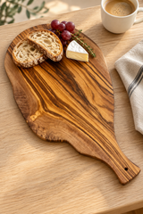 An olive wood serving board with a handle, styled with sliced bread, grapes, and a wedge of brie cheese on a light wood table next to a cup of coffee and a napkin.
