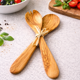 Wooden salad servers on a textured surface with a bowl of salad and ingredients in the background.