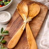 Two wooden spoons tied together on a wooden board with a salad in the background.