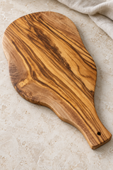 An olive wood serving board with a distinctive dark and light grain pattern, shown on a light beige stone surface with a neutral fabric napkin in the corner.