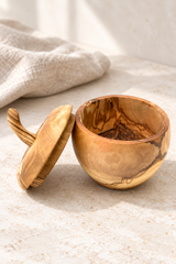 A small handcrafted apple shaped wooden bowl with its lid leaning beside it on a light stone surface. The bowl and lid have a warm honey-brown finish with natural darker wood grain patterns.
