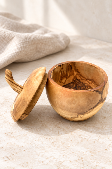 A small handcrafted apple shaped wooden bowl with its lid leaning beside it on a light stone surface. The bowl and lid have a warm honey-brown finish with natural darker wood grain patterns.