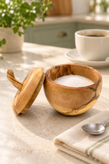 A small carved wooden bowl with its lid leaning beside it, filled with white sugar, set on a light stone countertop in a kitchen. A folded cream towel with a green stripe, a silver spoon, and a white cup of coffee are visible in the background.