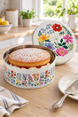 A round floral tin cake box with a powdered sponge cake and jam filling, shown on a wooden table with its lid leaning behind it in a bright kitchen setting.