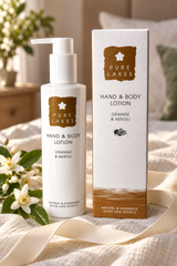 A bottle of Pure Lakes Hand & Body Lotion and its matching outer box, both labeled Orange & Neroli, styled on a cream fabric surface with white flowers and a soft indoor background.