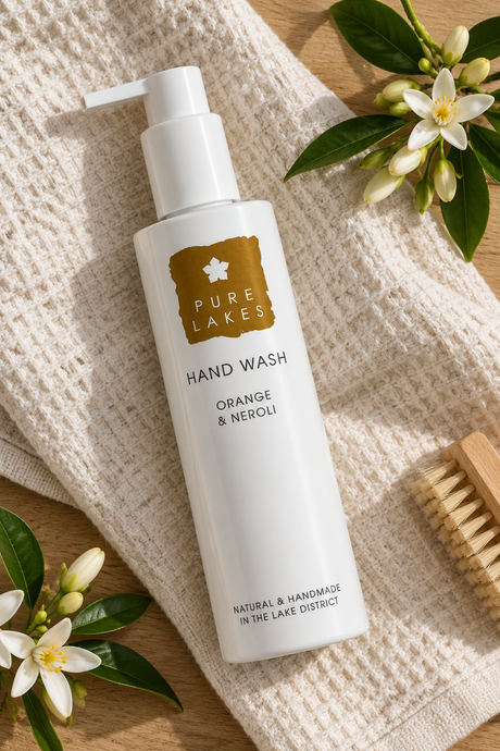 A bottle of Pure Lakes Hand Wash in Orange & Neroli scent, styled on a textured cream towel with orange blossoms, green leaves, and a wooden brush on a light wood surface.
