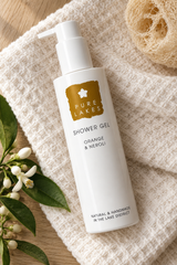 A bottle of Pure Lakes Shower Gel in Orange & Neroli scent, styled on a textured cream towel with green leaves, white blossoms, and a natural loofah sponge.