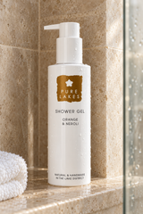 A bottle of Pure Lakes Shower Gel in Orange & Neroli scent, placed on a beige stone shower ledge with water droplets, next to a white towel.