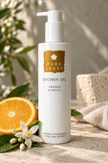 A bottle of Pure Lakes Shower Gel in Orange & Neroli scent, styled with a fresh orange slice, a white flower, and green leaves on a textured stone surface.
