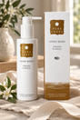 Pure Lakes Hand Wash bottle and matching box on a soft beige fabric surface with flowers and a vase in the background. The product is labeled Orange & Neroli.
