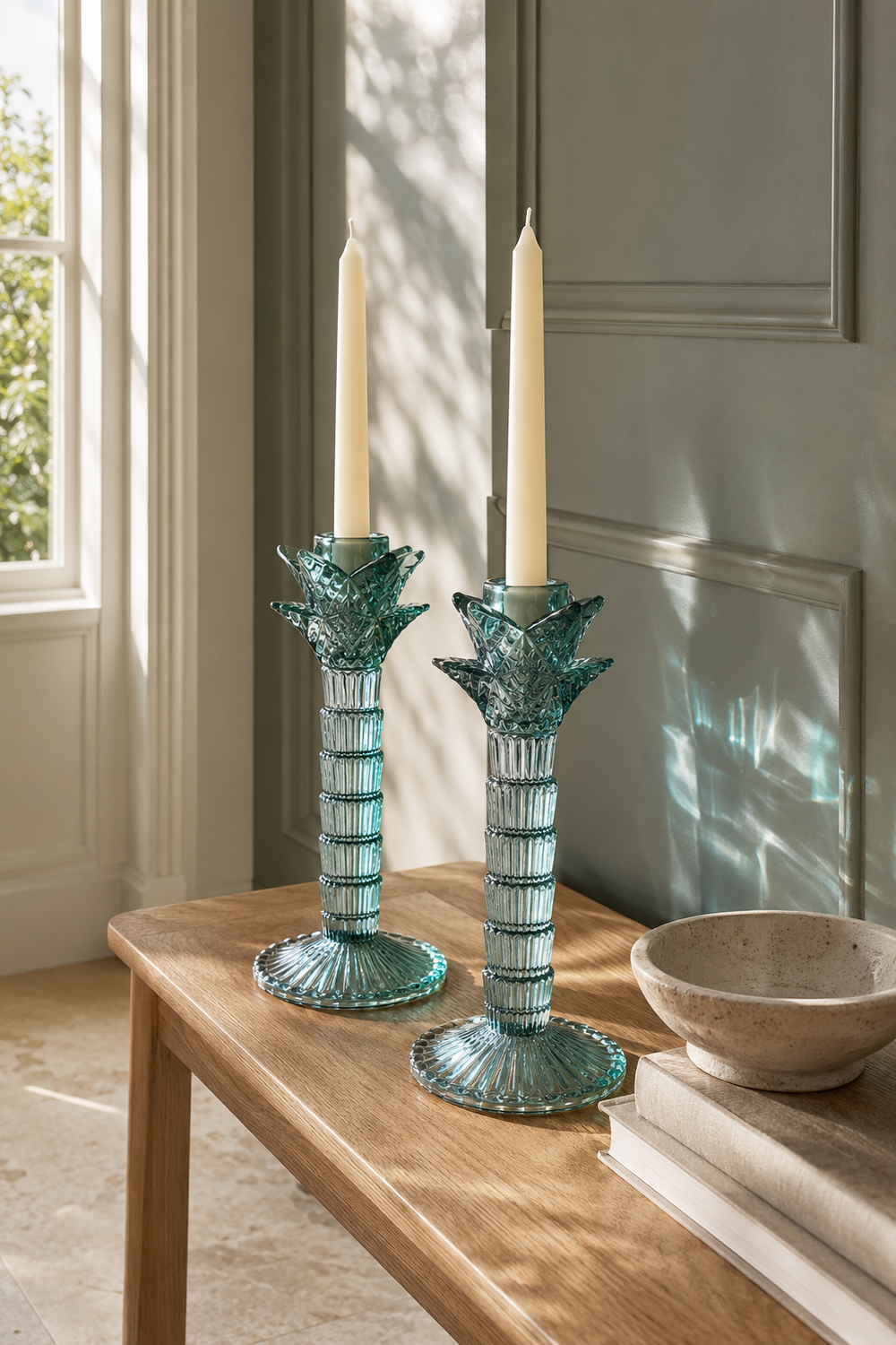 Two matching teal glass candlestick holders with fluted bases and leaf-like candle holders, each holding a white taper candle, displayed on a wooden table in a sunlit room.
