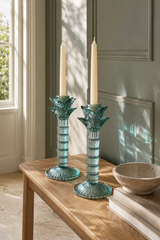 Two matching teal glass candlestick holders with fluted bases and leaf-like candle holders, each holding a white taper candle, displayed on a wooden table in a sunlit room.