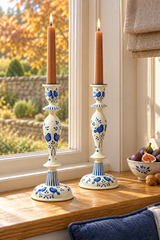 Two tall wood candlesticks with blue floral patterns and lit taper candles, displayed on a wooden windowsill in a bright room.